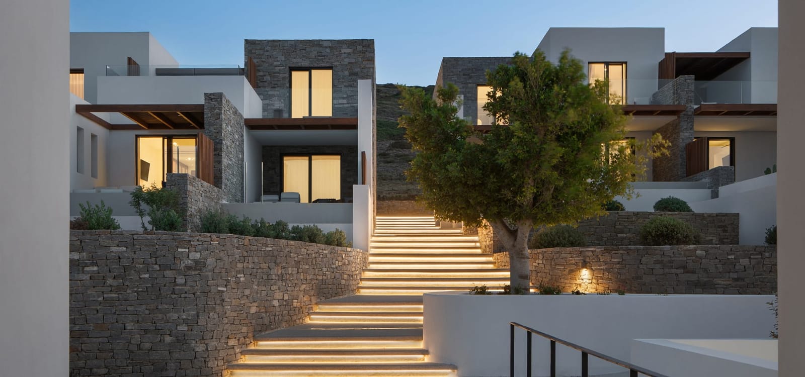 Stone-clad guest buildings of Hotel Vione and the lit stone stairway through the site at dusk